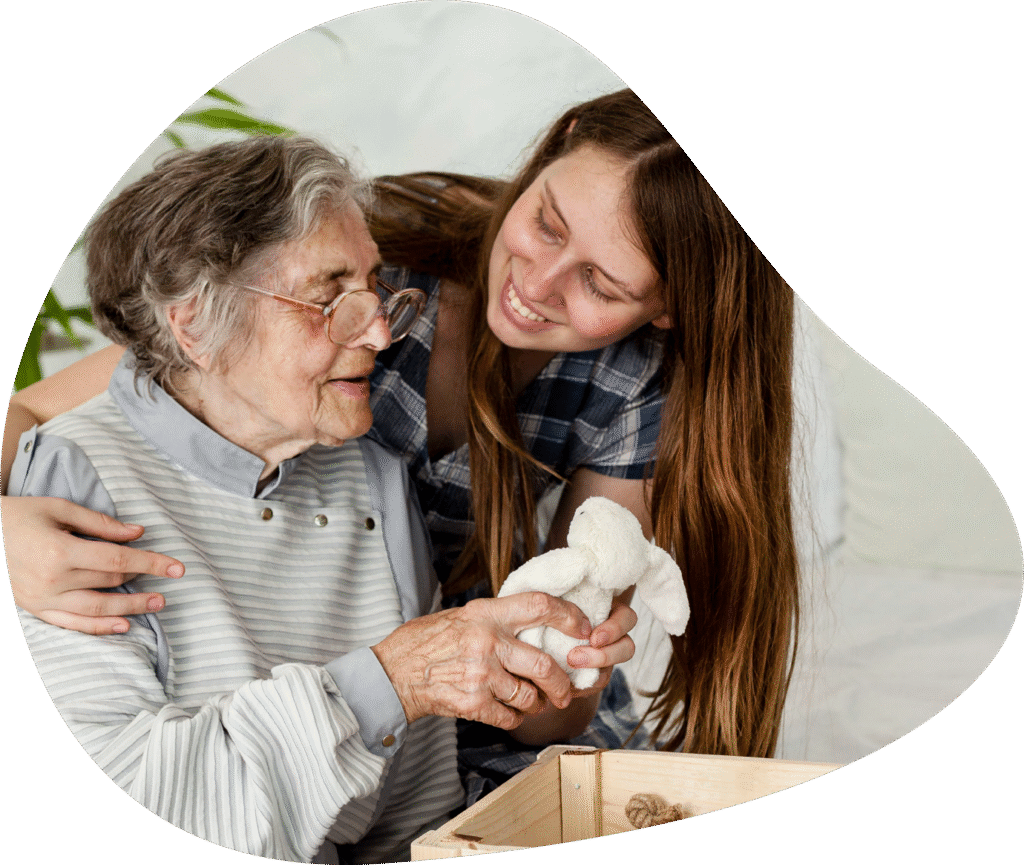 Young caregiver and elderly woman sharing joyful moment with stuffed animal during home care visit in Denver.